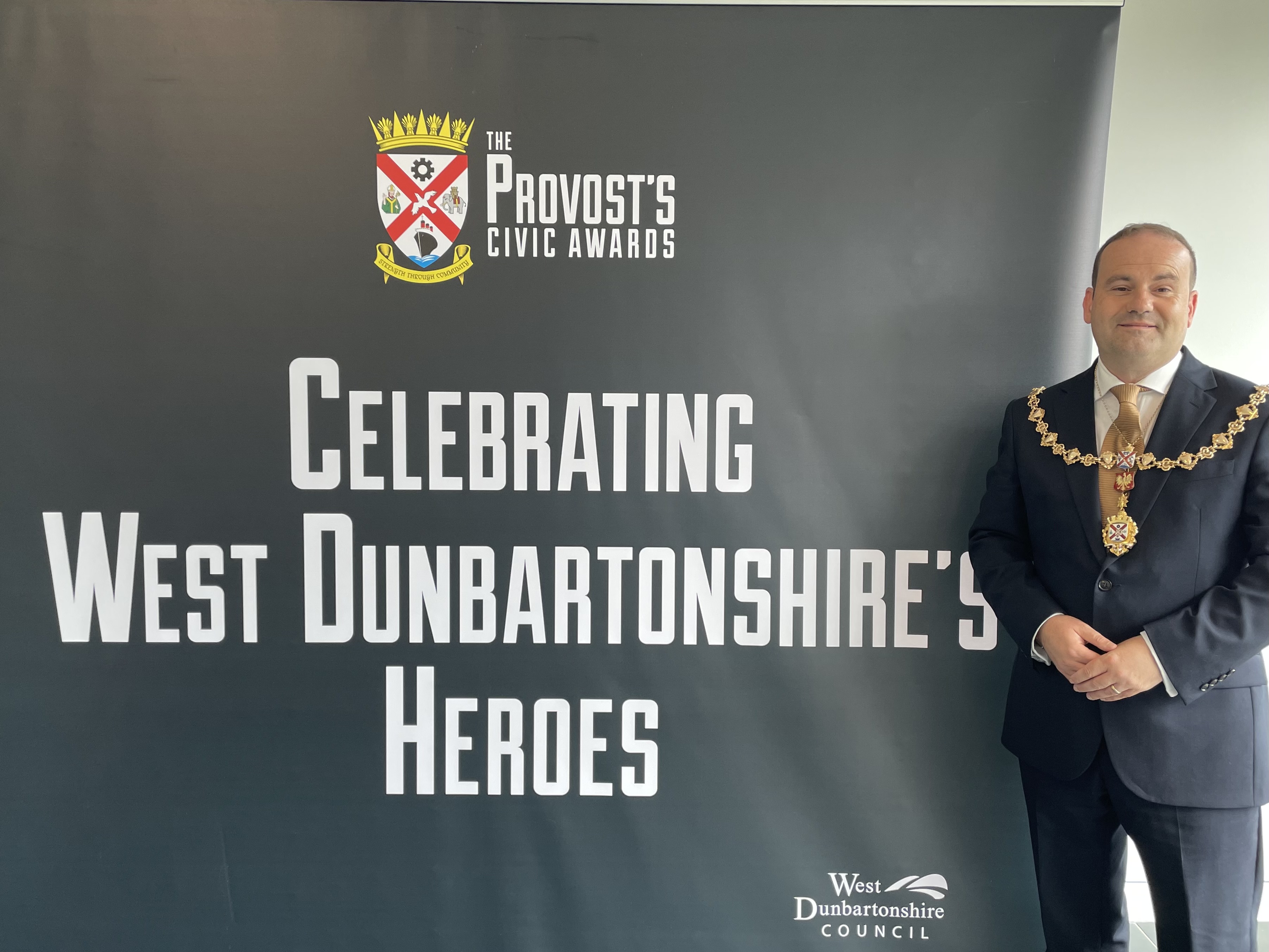 Provost Douglas McAllister in his Provost chains standing next to awards poster