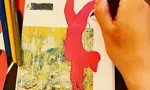 A close-up, top-down view of a person’s hand using a purple pencil to draw on a handmade zine. The zine features a collage of a person in a red outfit performing a handstand over an autumnal landscape. Coloured pens and paper scraps are scattered on the wooden table nearby.